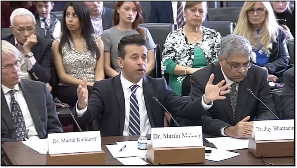 A man speaking emphatically with his hands raised during a conference or hearing, seated between two other individuals. Several people are seated in the background, listening attentively. Nameplates for Dr. Martin Kulldorff and Dr. Jay Bhattacharya are visible.