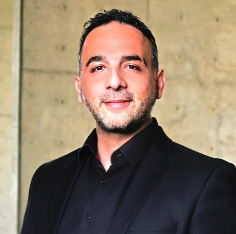 A man with short dark hair and a beard, wearing a black suit and black shirt, is smiling slightly while standing against a beige concrete wall.