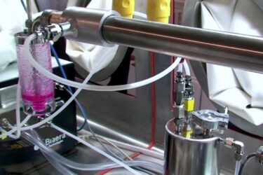 A laboratory apparatus featuring a series of connected stainless steel pipes and tubes with clear plastic tubing. The setup includes containers, one of which holds a pink liquid. Various clamps and connectors are visible on the equipment.