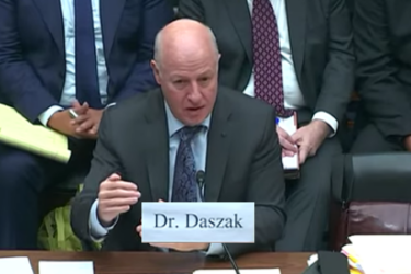 An individual labeled as Dr. Daszak is speaking at a formal meeting, seated at a table with microphones and documents. Several people in suits are sitting in the background.