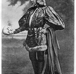 A black-and-white photo of a person dressed in an elaborate costume, standing and holding a skull in one hand. The backdrop has a cloudy sky. The style suggests a theatrical setting.