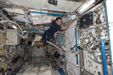 An astronaut floats inside the International Space Station, surrounded by equipment, cables, and storage bags. The setting appears to be a lab module, with various devices and panels on the walls and ceiling.