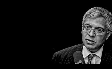 A grayscale image of a man wearing glasses and a suit, speaking into a microphone against a black background.