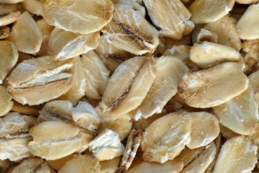 Close-up of raw, rolled oats displaying their flat, oval shape with beige and white shades. The texture appears dry and flaky, typical of uncooked oatmeal.