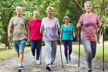 A group of five elderly people, three women and two men, walk on a path in a park. Two women are using Nordic walking poles. The group is smiling and wearing casual, sporty clothing. Trees and greenery surround the path.