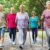 A group of five elderly people, three women and two men, walk on a path in a park. Two women are using Nordic walking poles. The group is smiling and wearing casual, sporty clothing. Trees and greenery surround the path.