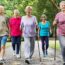 A group of five elderly people, three women and two men, walk on a path in a park. Two women are using Nordic walking poles. The group is smiling and wearing casual, sporty clothing. Trees and greenery surround the path.