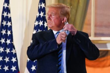 A man in a suit stands in front of two American flags, removing a face mask. He is looking to the side with a serious expression. The background shows a building with large windows.