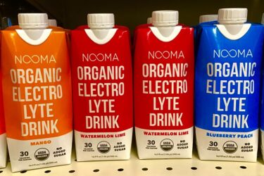 Four cartons of NOOMA Organic Electrolyte Drink are displayed: Mango (orange), Watermelon Lime (red), and two Blueberry Peach (blue). Each has "No Added Sugar" and USDA Organic labels, containing 30 calories per carton.