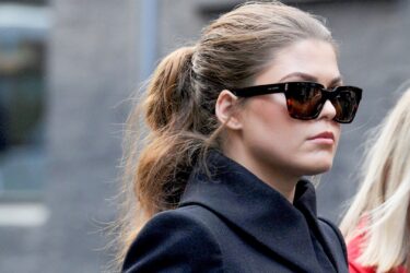 A woman with brown hair tied back wears large, dark sunglasses and a dark coat. She is walking outside, with a blurred urban background. Another person is partially visible beside her.