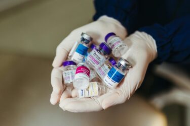 A person wearing white gloves holds several small Botox vials with purple, pink, and blue caps, labeled with various brands. The vials appear to contain injectable solutions.