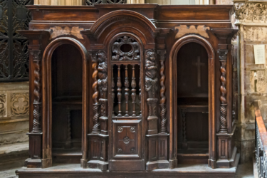 A traditional wooden confessional booth with ornate carvings and two entry arches, located in an ornate church setting. The central section is intricately detailed, featuring decorative columns and woodwork.