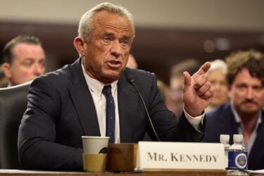 A man in a suit, identified as Mr. Kennedy, gestures while speaking at a microphone during a formal meeting or hearing. He has gray hair and is sitting at a table with name placard, papers, a coffee cup, and water bottles. People are visible in the background.