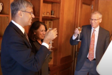 Three people in business attire are raising glasses for a toast. They are standing in a warmly lit room with wooden bookshelves in the background. Two men and one woman appear to be smiling and engaged in a positive interaction.