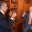 Three people in business attire are raising glasses for a toast. They are standing in a warmly lit room with wooden bookshelves in the background. Two men and one woman appear to be smiling and engaged in a positive interaction.