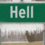 A green highway sign with the word "Hell" is covered in frost and icicles, suggesting a humorous contrast to the typical connotations of "Hell" being hot.