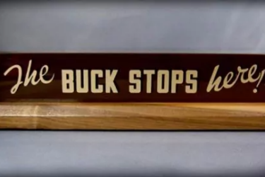 A wooden desk sign reads "The BUCK STOPS here!" in bold, white letters on a dark background. The sign sits on a light wood base against a neutral backdrop.