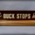 A wooden desk sign reads "The BUCK STOPS here!" in bold, white letters on a dark background. The sign sits on a light wood base against a neutral backdrop.