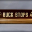 A wooden desk sign reads "The BUCK STOPS here!" in bold, white letters on a dark background. The sign sits on a light wood base against a neutral backdrop.