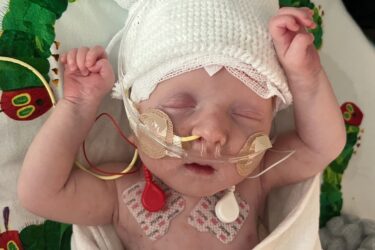 A newborn baby lies on a hospital bed with arms raised, wearing a white knit hat and connected to medical tubes and sensors. The baby is covered with a blanket, and part of a colorful caterpillar design is visible nearby.