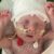 A newborn baby lies on a hospital bed with arms raised, wearing a white knit hat and connected to medical tubes and sensors. The baby is covered with a blanket, and part of a colorful caterpillar design is visible nearby.