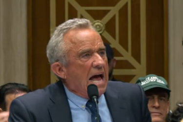 A man in a suit, labeled as "Secretary KENNEDY," speaks passionately into a microphone at a hearing. He sits between two men in suits, with an audience, cameras, and microphones visible in the background.