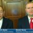 Dr. Mark Geier and David Geier, both in suits, sit side by side during a televised interview. Bookshelves and a wooden door are behind them, while the FOX19 NOW logo and text appear at the bottom of the screen.