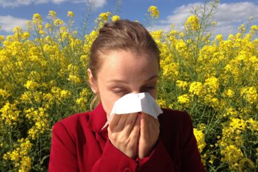 A woman in a red jacket holds a tissue to her nose, appearing to sneeze or blow her nose, while standing in front of a field of yellow flowers under a blue sky with scattered clouds.