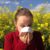 A woman in a red jacket holds a tissue to her nose, appearing to sneeze or blow her nose, while standing in front of a field of yellow flowers under a blue sky with scattered clouds.