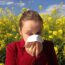 A woman in a red jacket holds a tissue to her nose, appearing to sneeze or blow her nose, while standing in front of a field of yellow flowers under a blue sky with scattered clouds.