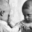 A baby wearing a long-sleeved shirt touches a mirror, looking intently at its own reflection while pointing at the glass with one finger.