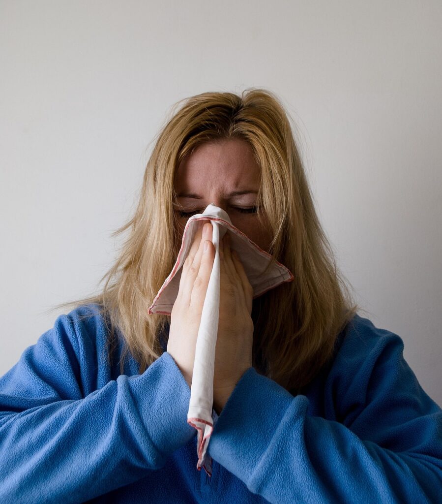 A person with blonde hair wearing a blue sweater is holding a handkerchief to their nose, appearing to sneeze or blow their nose against a plain, light-colored background.
