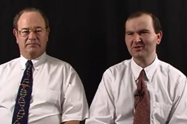 Two men in white shirts and patterned ties sit side by side against a plain black background, looking toward the camera.