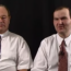 Two men in white shirts and patterned ties sit side by side against a plain black background, looking toward the camera.