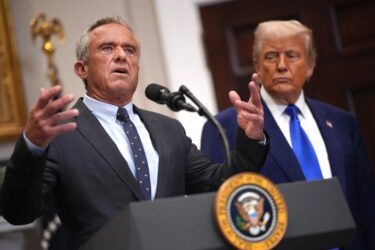 A man in a suit speaks at a podium with the U.S. presidential seal, gesturing with his hands, while another man in a suit stands behind him, listening attentively.