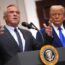 A man in a suit speaks at a podium with the U.S. presidential seal, gesturing with his hands, while another man in a suit stands behind him, listening attentively.