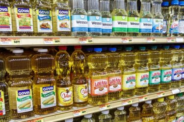 Bottles of various vegetable oils, including brands like Goya, Wesson, LouAna, and Crisco, are neatly arranged on grocery store shelves. Price tags are visible below the rows of oils.