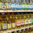 Bottles of various vegetable oils, including brands like Goya, Wesson, LouAna, and Crisco, are neatly arranged on grocery store shelves. Price tags are visible below the rows of oils.