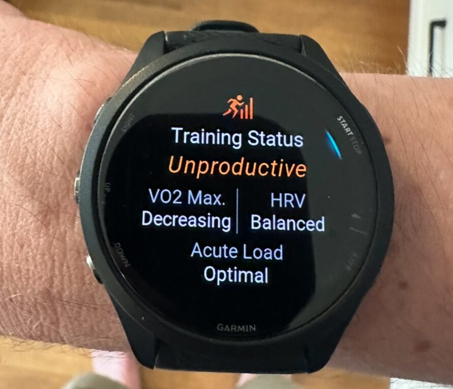 A smartwatch on a person's wrist displays training status as "Unproductive," with VO2 Max decreasing, HRV balanced, and acute load optimal. The watch has a black band and is on a light-skinned arm.