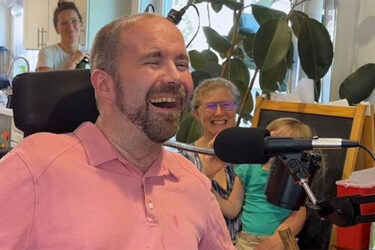 A man in a pink shirt smiles while speaking into a microphone. In the background, two women and a young child sit and smile in a bright, homey room with plants and kitchen items visible.
