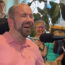 A man in a pink shirt smiles while speaking into a microphone. In the background, two women and a young child sit and smile in a bright, homey room with plants and kitchen items visible.