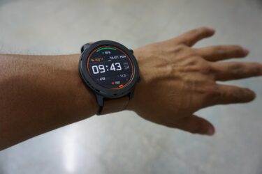 A person’s left wrist wearing a round black smartwatch displaying the time 09:43, date, battery level, heart rate, steps, and other fitness data on its colorful digital screen.