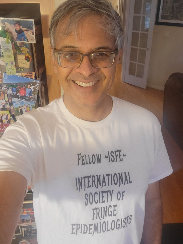 A smiling person with gray hair and glasses wears a white T-shirt that reads, "Fellow ~ISFE~ International Society of Fringe Epidemiologists." They are standing indoors in a well-lit room.