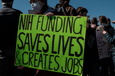 A person in a crowd holds a bright green sign that reads, "NIH FUNDING SAVES LIVES & CREATES JOBS." Other people are visible around, some wearing jackets and one wearing a mask.
