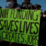 A person in a crowd holds a bright green sign that reads, "NIH FUNDING SAVES LIVES & CREATES JOBS." Other people are visible around, some wearing jackets and one wearing a mask.