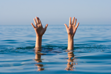 Two hands are raised above the surface of a body of water, suggesting a person is struggling or in distress, possibly drowning, with a clear blue sky and calm water in the background.