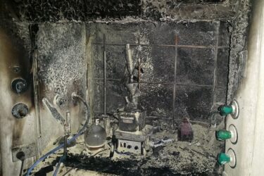 A laboratory fume hood heavily damaged by fire, with charred equipment, melted glassware, and black soot covering the surfaces; burn marks and debris are visible throughout the scene.