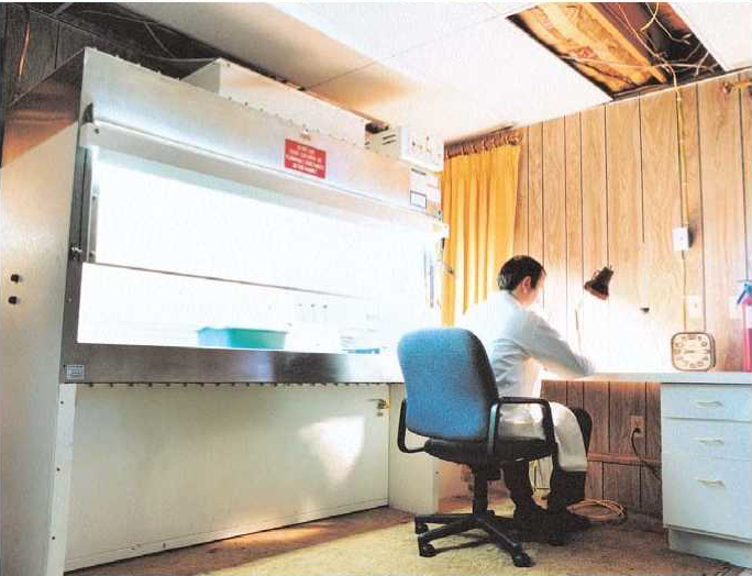 A person in a lab coat sits at a desk in a small, dimly lit room with wood-paneled walls, working under a desk lamp next to a large laboratory fume hood. The ceiling above shows exposed pipes.