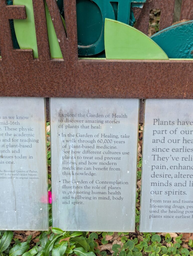 A metal sign reads: "Explore the Garden of Health to discover amazing stories of plants that heal," followed by descriptions of the Garden of Healing and the Garden of Contemplation’s roles in medicine and wellbeing.