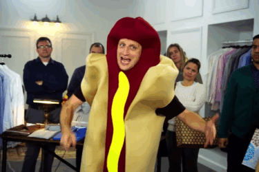A man in a hot dog costume gestures animatedly while standing in front of a group of people in an office setting, some of whom appear amused or bemused.